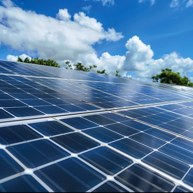 Großaufnahme von Photovoltaik-Solarmodulen unter einem klaren blauen Himmel mit vereinzelten weißen Wolken. Das Bild zeigt die glänzenden Oberflächen der Solarpaneele, die die Sonnenstrahlen reflektieren und die Nutzung erneuerbarer Energiequellen hervorheben. Im Hintergrund sind Bäume zu sehen, was die Harmonie zwischen moderner Solartechnologie und der Natur betont. Diese Solaranlage steht symbolisch für nachhaltige Energiegewinnung und Umweltschutz.